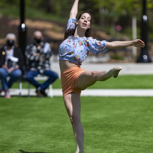 Single dancer in flowered shirt in a dance pose with leg extended on green grass, outdoors